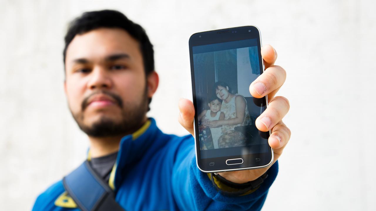 Miguel Flores holding a phone that displays a photo of Miguel with his mom at a younger age