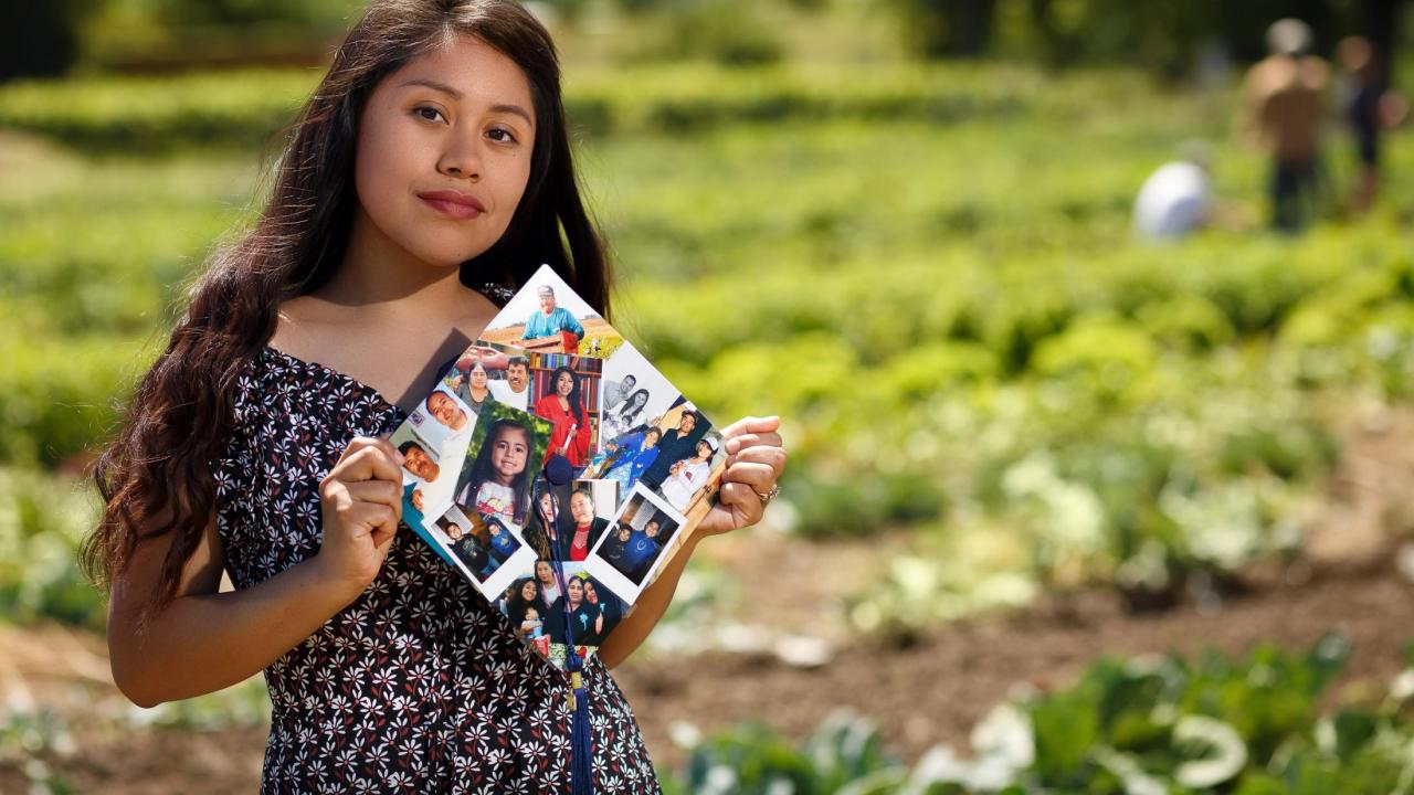 First-generation student, Yilda Korepela standing in the UC Davis student farm holding her graduation cap that features photos from her family and childhood. 