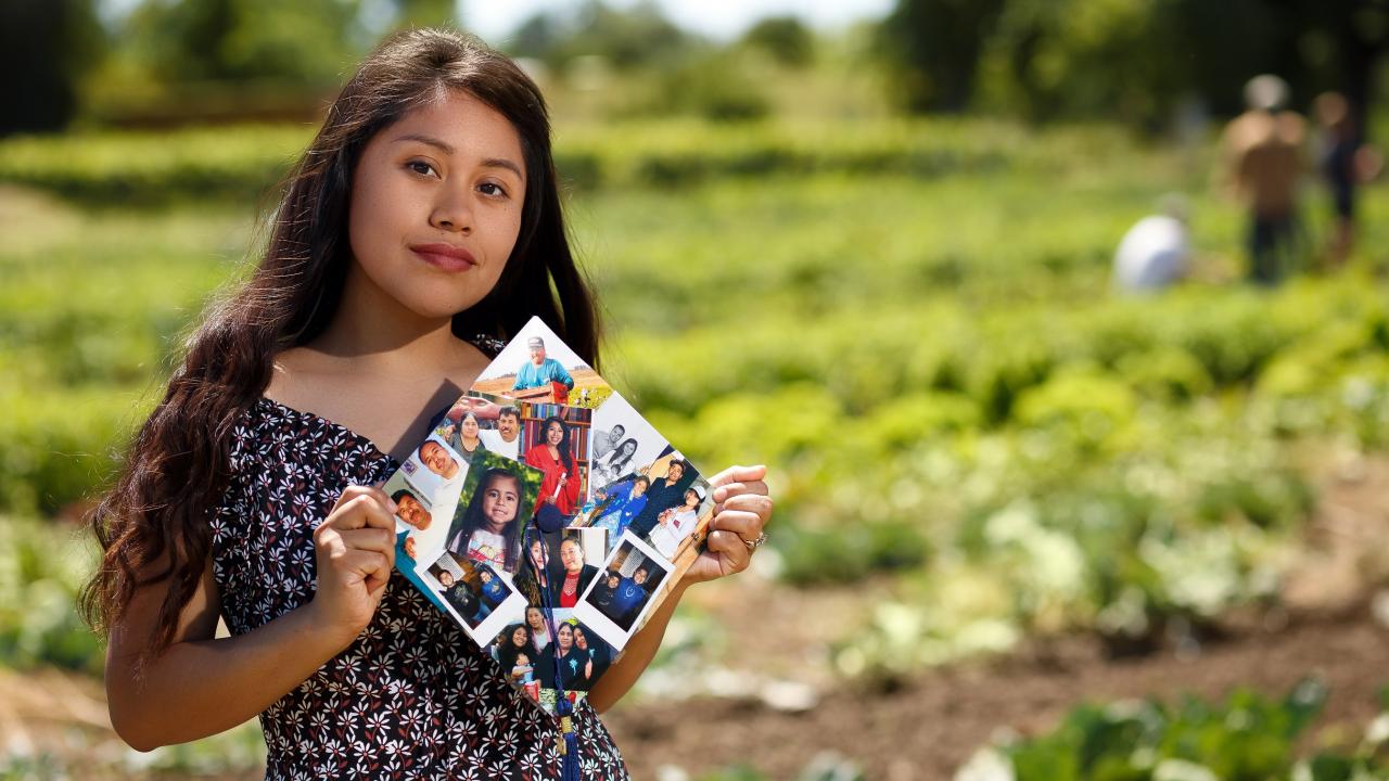 First-generation student, Yilda Korepela standing in the UC Davis student farm holding her graduation cap that features photos from her family and childhood. 