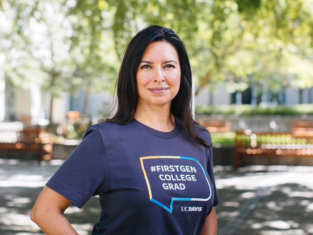 Jeanette B. Ruiz stands with a hand on her hip in the middle of Shields Library courtyard. She is wearing a blue T-shirt with a colorful graphic that says #firstgen college grad UC Davis.